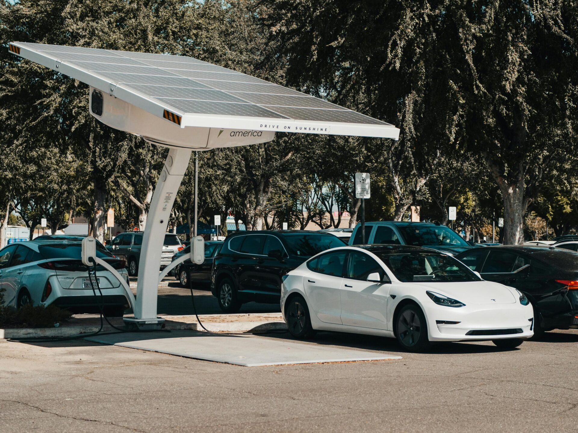 A white Tesla Model 3 is parked and charging at a solar-powered EV charging station in a parking lot. The station features a large canopy with solar panels and the phrase “Drive on Sunshine” printed on the side. Other parked vehicles and trees are visible in the background, suggesting a sustainable energy setting.