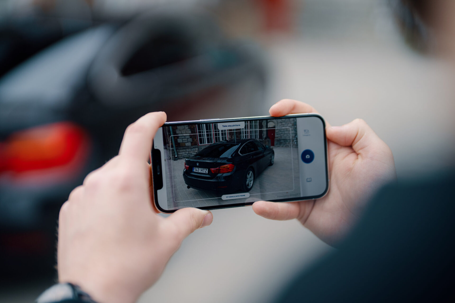 Close-up of a person using a smartphone to take a guided photo of a black car with the DriveX app, as part of a digital vehicle inspection. The phone screen displays the car within an on-screen frame.