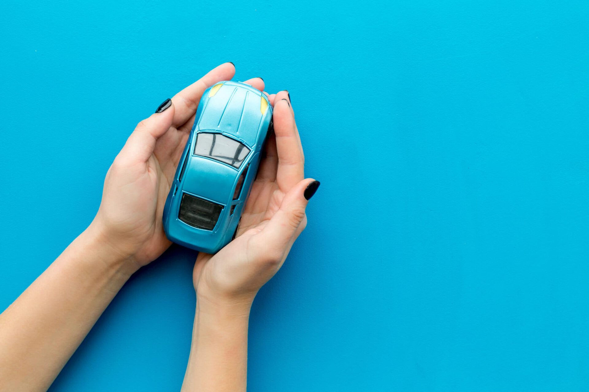 A pair of hands with black-painted fingernails gently holding a small blue toy car against a solid blue background. The hands are positioned to cradle the car protectively, creating a visual metaphor for car care, safety and insurance.
