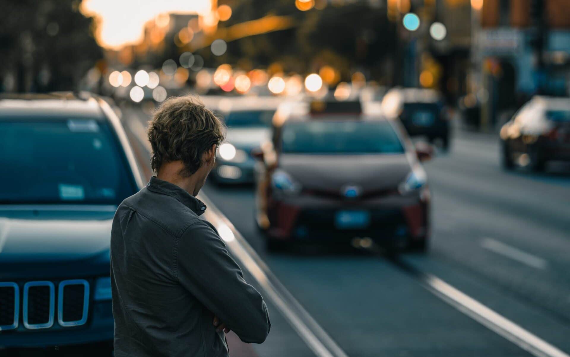Man standing beside a parked car, looking toward oncoming traffic on a busy city street at sunset.