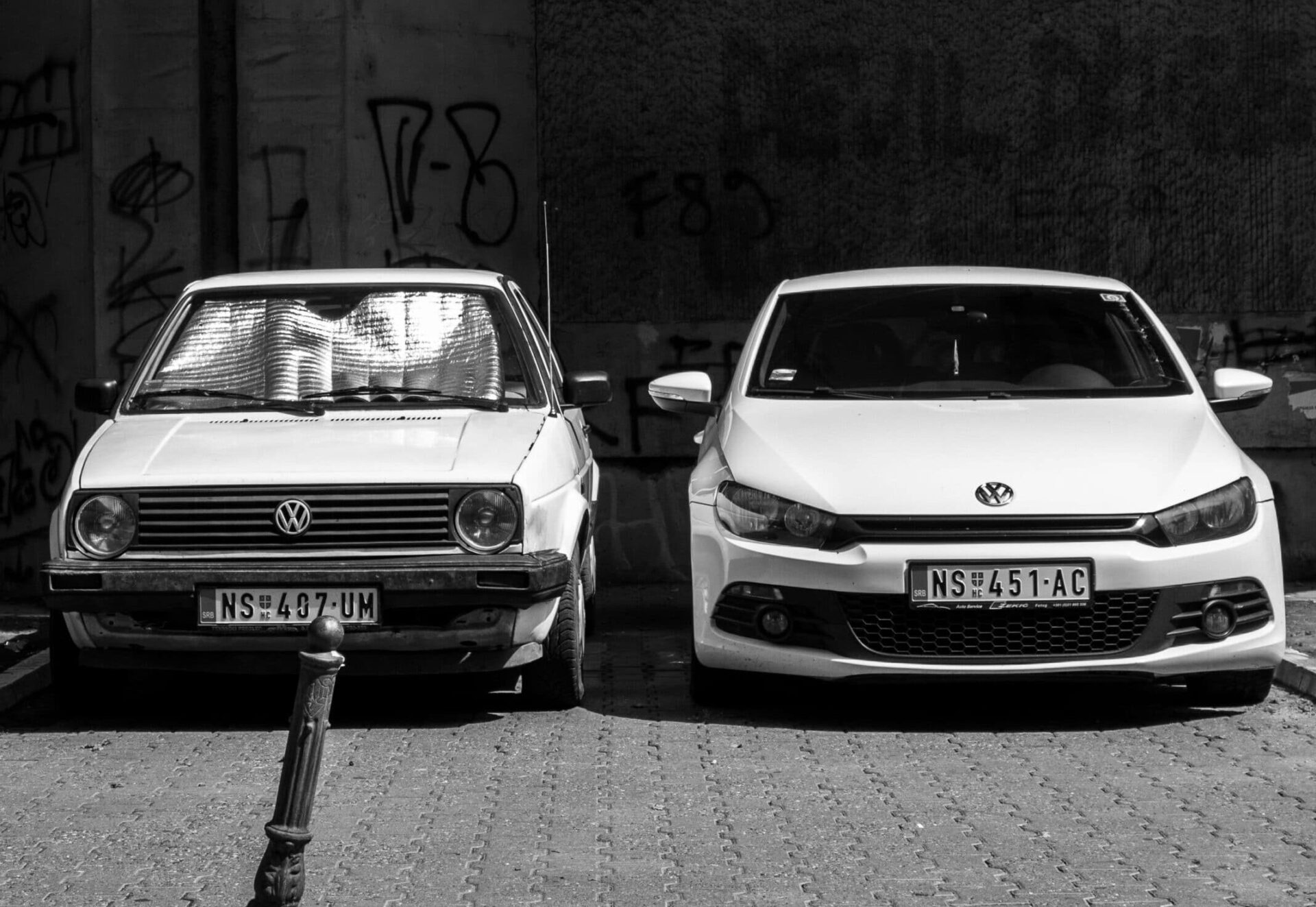 A black and white photograph of two Volkswagen cars parked side by side in front of a graffiti-covered wall. On the left is an older, worn-out model with a reflective sunshade in the windshield and visible damage. On the right is a newer, sleek model in better condition. Both cars have Serbian license plates starting with “NS.” The contrast highlights the evolution in car design over time.