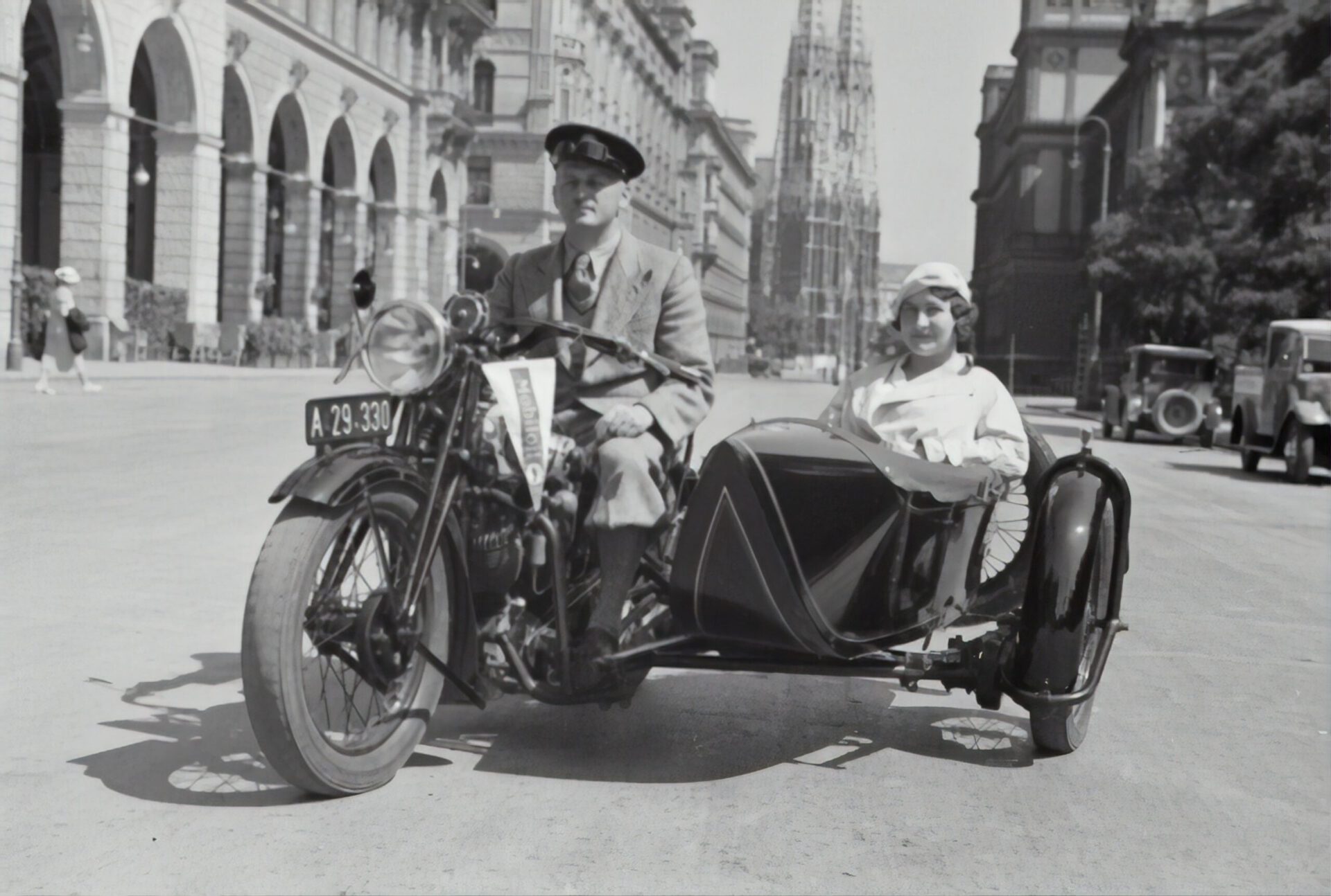 A black and white photograph of a man driving a vintage motorcycle with a sidecar carrying a smiling woman. They are on a wide city street with historic European architecture, including arched colonnades and a tall, ornate cathedral tower in the background. Several early 20th-century cars are parked along the street. The man wears a suit and cap, while the woman is dressed in light-colored attire and a headscarf.
