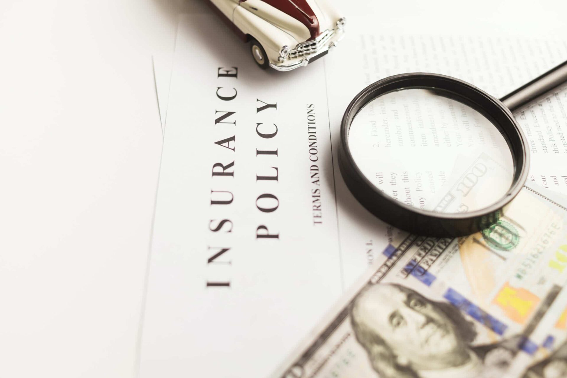 Close-up of an insurance policy document with a toy car, magnifying glass, and US $100 bill, symbolizing car insurance and financial coverage.