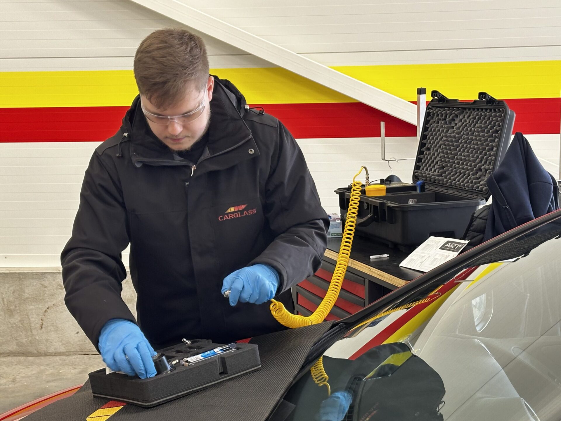 A technician wearing a black Carglass-branded jacket, safety goggles, and blue gloves is preparing tools for a windshield repair. He is working at the front of a vehicle with a specialized repair kit on the hood. In the background, a black equipment case with foam padding, a yellow coiled air hose, and a workbench are visible against a white wall with red and yellow horizontal stripes.